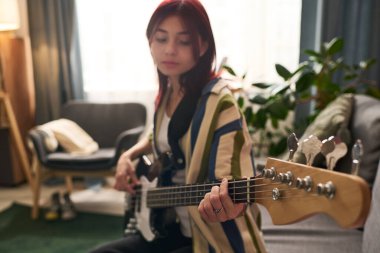 Arka planda rahat mobilyalar ve bitkilerle rahat bir evde otururken bas gitar çalan bir kız. Oda doğal ışıkla dolu. Sıcak atmosfer yaratıyor.