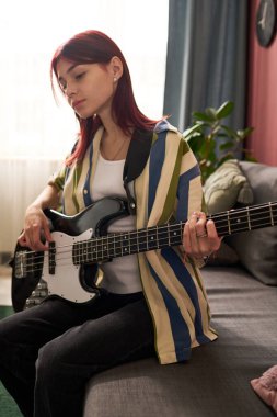 Rahat bir oturma odasında kanepede oturmuş gitar çalan ve camdan içeri ışık süzülen bir kız.