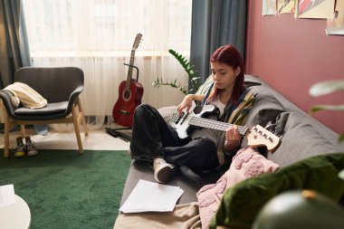 Genç kız kanepede oturuyor ve rahat oturma odasında yere dağılmış müzik aletleri ve kağıtlarla birlikte elektro gitar çalıyor. Pencereden içeri gün ışığı giriyor