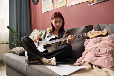 Kanepede oturmuş gitar çalan kızıl saçlı genç bir kız. Arka planda kırmızı duvarda dekoratif posterler ve rahat atmosfer için yumuşak yastıklar yer alıyor