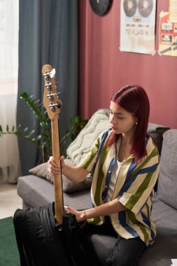 Boyalı kızıl saçlı genç kız rahat bir odada, elinde elektro gitarla oturuyor, duvarlarda posterler asılı, arka planda ev bitkisi var.