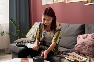 Kanepede oturan kız hoparlör kullanırken elektro gitar çalıyor. Rahat yastıklar ve bitkilerle dekore edilmiş bir oda rahatlatıcı bir atmosfer yaratıyor.