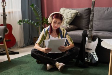 Genç bir kız yerde oturmuş doküman okurken kulaklıkla arkasında gitar ve saksı bitkisi olan gri bir kanepe dinliyor.