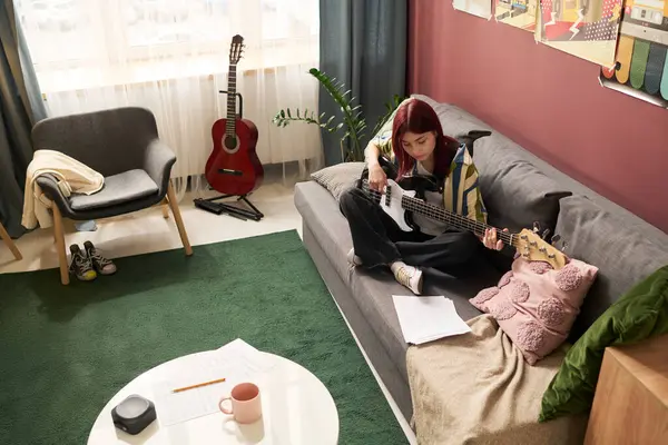 Gri kanepede oturan genç kız pencere kenarındaki odada yeşil halı, yuvarlak masa, sandalye, kitap ve bitki içeren akustik gitar çalıyor.