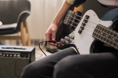 Konforlu oturma odasında elektro bas gitar çalan kişilerin yakın çekimleri ve arka planda yükseltici teçhizatı ve sandalye ile müzik çalışması için davetiye alanı yaratmak