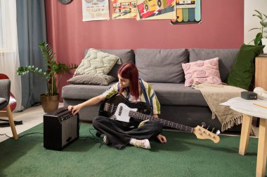 Kızıl saçlı genç kız oturma odasında modern dekorlu ve yeşil halılı elektrikli bas gitar akort ediyor. Müzik meraklısı amplifikatör ayarlarını ayarlıyor