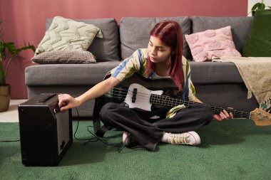 Genç kız, arka planda kanepede çiçek ve yastıklarla rahat oturma odasında amplifikatöre bağlı olarak bas gitar çalıyor.