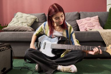 Oturma odasında yerde otururken bas gitar çalan bir kız. Yastıklarla çevrili ve yakınlarda yükseltici olan rahat bir ortam var.