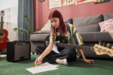Genç kız yerde elektro gitarla oturuyor, kağıda notalar yazıyor. Etrafı müzik ekipmanları ve posterlerle çevrili, rahat uzayda sanat yaratıyorlar.