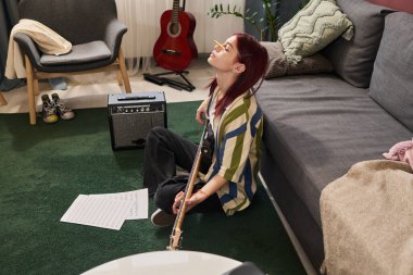 Oturma odasında akustik gitar tıngırdatırken halıda oturan genç bir adam. Yanında müzik ve müzik ekipmanları var. Duvara yaslanmış kırmızı elektro gitar