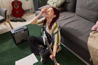 Yeşil halıda oturan, elektro gitar çalan ve etrafında müzik ekipmanları ve rahat bir oturma odasında gitar çalan bir kız.