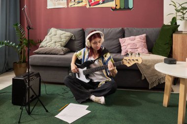 Yerde oturan bir kız amplifikatör ve notalarla birlikte elektro bas gitar çalıyor. Rahat oturma odasında renkli bir dekor ile çevrili. Müzik yaratıcılığını teşvik etmek