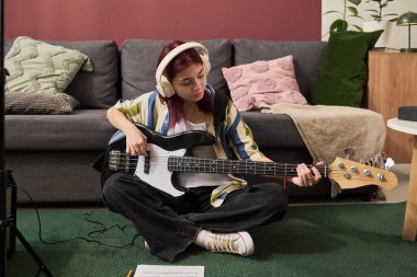 Oturma odasında halı döşenmiş yerde otururken ve rahat mobilyalarla çevrili kulaklıklı kız bas gitar çalıyor.