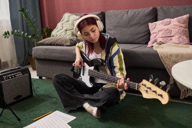 Genç bir kız, rahat bir oturma odasında yerde oturmuş, kulaklık takmış ve arka planda kanepe ve amplifikatörlerle notalara odaklanmış bir şekilde elektro gitar çalıyor.