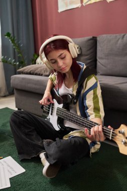 Genç beyaz kız yerde oturmuş, kulaklıkla bas gitar çalıyor. Etrafı kanepe ve iç mekan bitkileriyle çevrili. Rahatlatıcı bir ambiyans ekliyor.