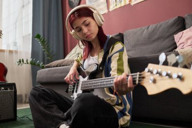 Rahat oturma odasında kulaklık takıp elektro gitar çalan genç bir kız. Odaklanmış ve rahatlamış hissediyor. Kanepe, dekor ve ev bitkileriyle çevrili.