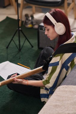 Kırmızı kulaklıklı genç kız kalem ve kağıdıyla yerde ders çalışıyor. Çevrede sandalye, masa ve odaklanmış bir ortam yaratan diğer çalışma materyalleri yer alıyor