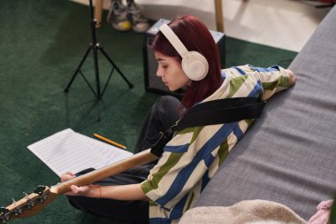 Genç kız yerde kulaklık takmış, bas gitar çalıyor ve nota okuyor. Amplifikatöre bağlı, rahatlatıcı bir atmosfer yaratıyor.