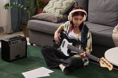 Genç bir kız yerde oturmuş bas gitar çalıyor kulaklıkla müzik dinliyor yanında yükseltici ve yeşil halıda nota kağıdı var.