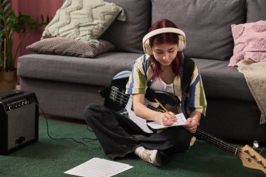 Genç kız yerde elektro gitarla oturuyor, not defterine şarkı sözü yazıyor. Müzik ekipmanlarıyla çevrili, kulaklık takan, şarkı yapımına odaklanmış ve dalmış gibi görünüyor.