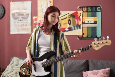 Kızıl saçlı genç bir kız. Müzik posterleriyle süslenmiş rahat bir odada gitar çalıyor. Ciddi ifadesine ve davranışlarına odaklan.