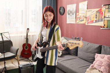 Konforlu bir oturma odasında gülümseyen ve duvarda müzik aletleri ve posterleri sergileyen bir kız, sıcak ve yaratıcı bir atmosfer yaratıyor.