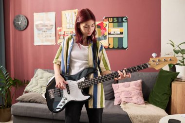 Genç kız rahat oturma odasında bas gitar çalıyor. Müzik posterleri duvarları süslerken yemyeşil bitki rahat uzaya biraz doğa katıyor.