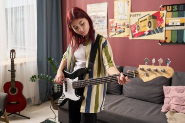 Genç kız rahat oturma odasında elektro gitar çalarken konsantre oluyor. Duvarlarında müzik posterleri ve akustik gitarları var.
