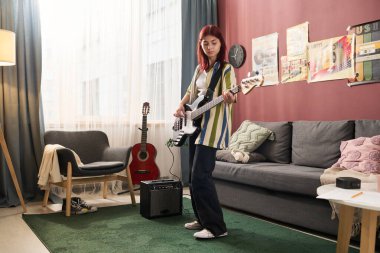 Çağdaş oturma odasında gitar çalan genç bir kız. Geniş bir iç mekan etrafında gitar yükseltici ve dekor elementleriyle rahat bir atmosfer yaratıyor.