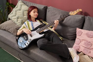 Bas gitar çalarken kanepede uzanan genç kız, gözleri odakta kapalı. Rahat yastıklı rahat ortam, arka planda yeşillik ve kızıl saç rengi.