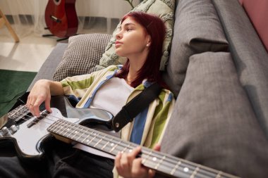 Genç bir kız, elinde elektro gitarla kanepeye yaslanıyor. Düşüncelere dalmış, arka planda müzik ekipmanları ve duvara yaslanmış akustik gitar da var.
