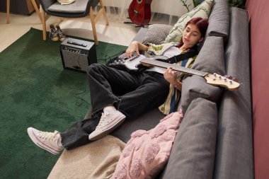 Genç insan, müzik ekipmanlarıyla çevrili bir şekilde, kanepede oturup bas gitar çalarak rahat bir hayat sürüyor.