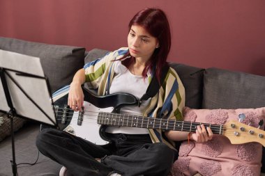 Önünde müzik sehpası olan bir kanepede otururken elektro bas gitar çalışmasına odaklanan kızıl saçlı kız. Yumuşak yastıklar ve sıcak renklerle günlük ev ortamları