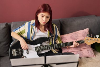 Kanepede oturup nota okurken bas gitar çalan kızıl saçlı kız. Yumuşak yastıklar ve sıcak duvar renkleri rahat bir atmosfer yaratır.