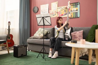 Oturma odasında amplifikatör, müzik standı ve arka planda kanepe ile otururken elektro gitar tellerini ayarlayan genç kız, duvardaki sanat eserleri ambiyansı yükseltiyor.