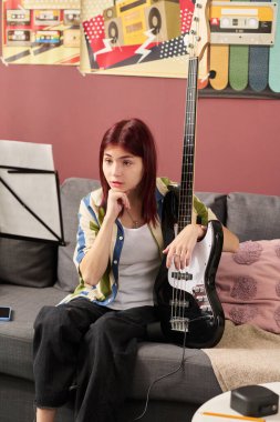 Kanepede oturan bir kızın portresi. Elinde elektro gitar. Düşüncelere dalmış, etrafı müzik ekipmanları ve dekorlarla çevrili.