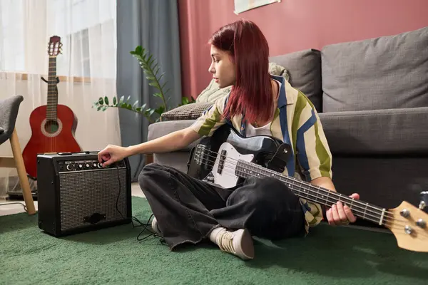 Halıya oturan kişi, elektro gitarı tutarken amplifikatör kontrollerini ayarlıyor, arka planda akustik gitar ve ev bitkisi, rahat bir yaşam alanı sergiliyor