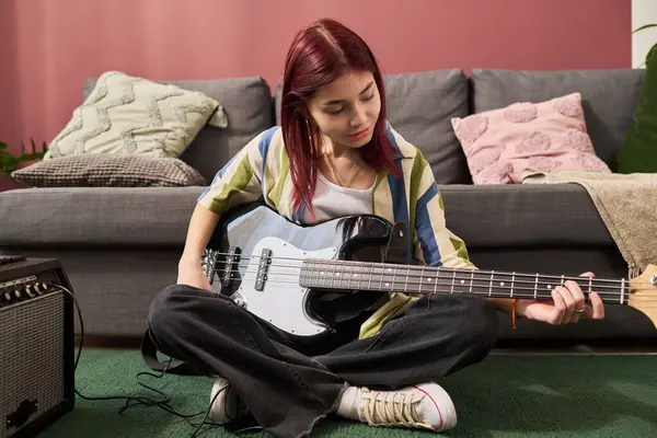 Oturma odasında yerde otururken bas gitar çalan bir kız. Yastıklarla çevrili ve yakınlarda yükseltici olan rahat bir ortam var.