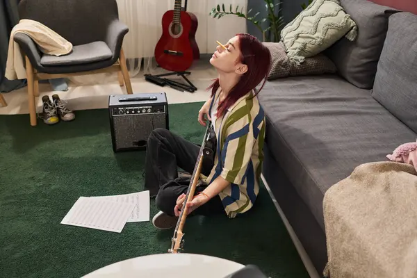 Oturma odasında akustik gitar tıngırdatırken halıda oturan genç bir adam. Yanında müzik ve müzik ekipmanları var. Duvara yaslanmış kırmızı elektro gitar