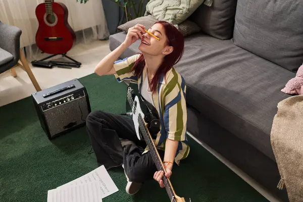 Yeşil halıda oturan, elektro gitar çalan ve etrafında müzik ekipmanları ve rahat bir oturma odasında gitar çalan bir kız.