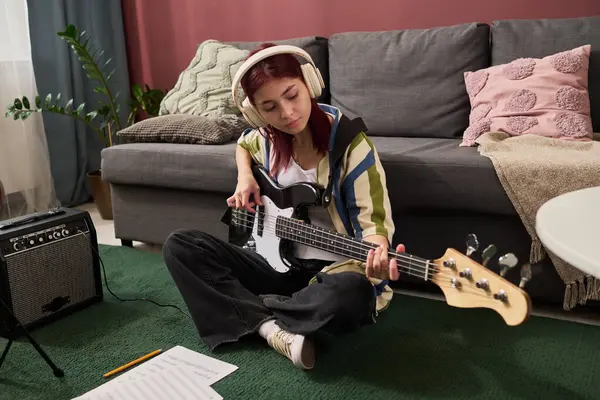 Genç bir kız, rahat bir oturma odasında yerde oturmuş, kulaklık takmış ve arka planda kanepe ve amplifikatörlerle notalara odaklanmış bir şekilde elektro gitar çalıyor.