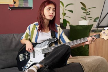 Genç kız kanepede oturuyor ve elinde düşünceli bir ifadeyle elektro gitarla arka planda saksı bitkisi ve renkli duvar sanatının kollarını kollarında dinlendiriyor.