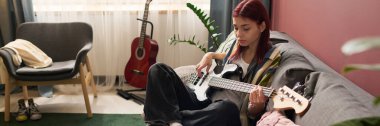 Kanepede oturan genç kız elektro gitar çalıyor, rahat oturma odasında rahat bir atmosfer yaratıyor, arka planda gitar ve yumuşak doğal ışıklandırma var.