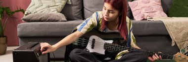 Kız, elinde bas gitarla salonda amfiyi ayarlarken yerde oturuyor. Kanepe ve yastıklar da dahil rahat bir ortam.