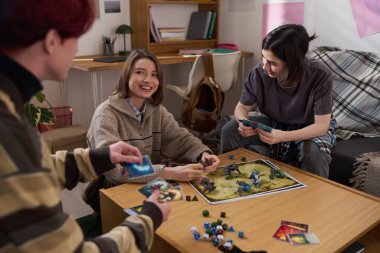 Yurt odasında arkadaşlarıyla masa oyunu oynayan ve üniversitede birlikte eğlenen genç bir kadının portresi.