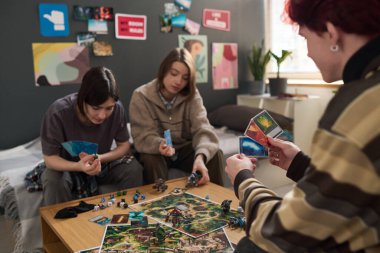 Üç gülümseyen gencin birlikte masa oyunu oynayıp kart ve minyatür oyun parçaları tutarak iletişim kurdukları gizli bir sahne.