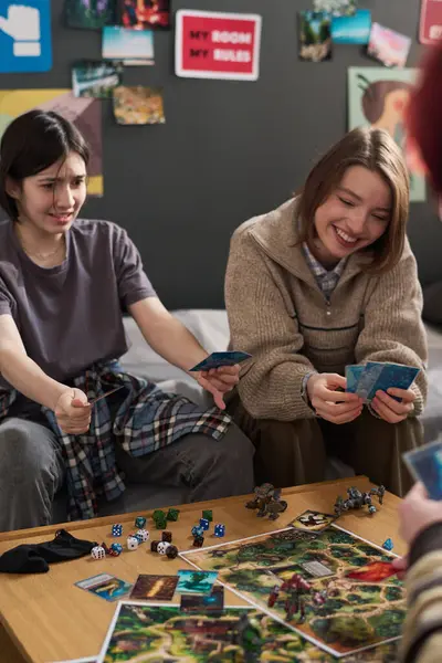 Evde ya da okulda oyun oynayan üç gencin dikey görüntüsü. Elinde kart tutan ve arkadaşlarıyla eğlenen gülen kızlara odaklan.