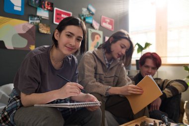 Oyun masası oturumunda arkadaşlarıyla rahat yurt ortamında resim çizerken kameraya bakan gülümseyen genç kızın portresi