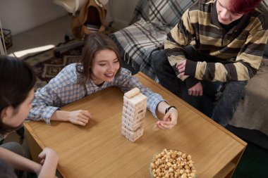Yatakhane ortamında arkadaşlarıyla masa oyunu oynarken tahta kuleden blok çeken gülümseyen genç kızı yüksek açıdan görmek.