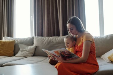 Oturma odasında, kanepede oturup kitap okuyan anne ve çocuğun yan görünüm portresi.